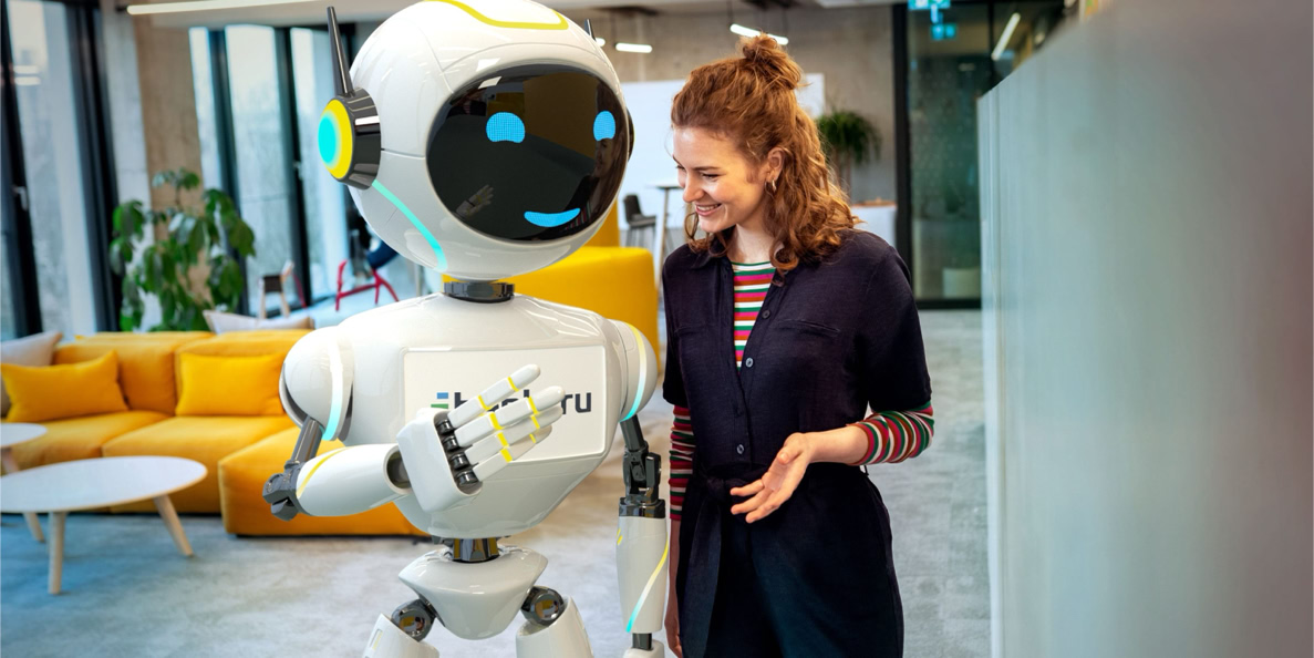 A woman smiling at a humanoid robot in an office setting with yellow couches and large windows in the background.