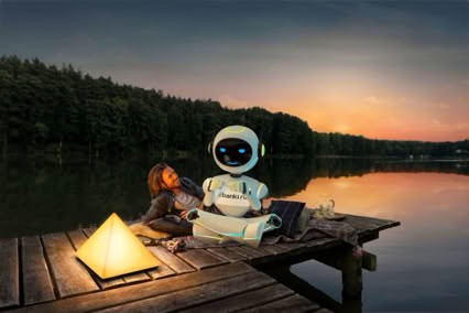 A person and a robot sitting on a wooden dock by a lake at sunset, with a glowing pyramid-shaped lamp beside them.